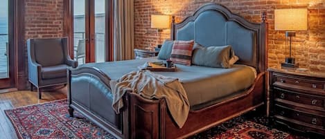 A bedroom with a large bed, a chair, a nightstand, and a view of the outdoors.