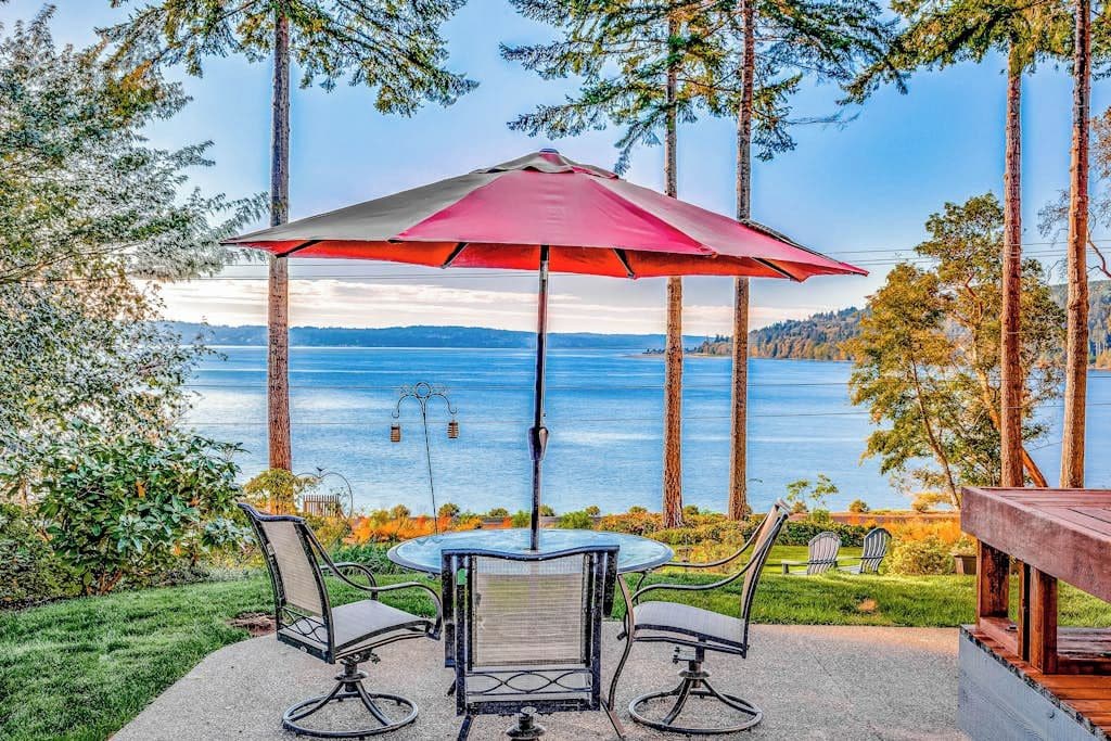 Un patio al aire libre con una mesa y sillas, un paraguas rojo y vistas a un lago.