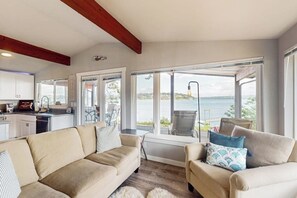 A modern living room with a sofa and armchairs, large windows with a view of the sea, and a kitchen in the background. - 2BR family friendly getaway with deck, Washer Dryer, Wifi (Fox Island)