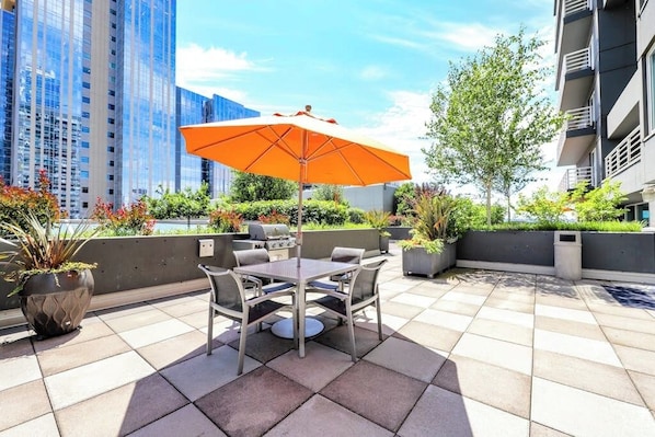 An outdoor patio with a table and chairs, surrounded by potted plants and a large orange umbrella. - 1BR condo near attractions with pool & hot tub (Seattle)