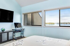 A bedroom with a bed, a desk with a computer, a TV, and a window with a view of a lake and trees.