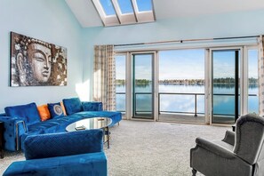 A modern living room with a large window overlooking a lake, a blue sofa set, and a Buddha painting on the wall.
