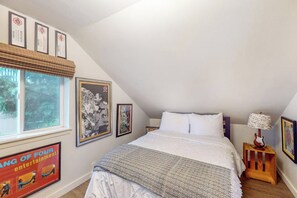 A bedroom with a sloped ceiling, a bed with white bedding, a wooden nightstand with a lamp, and framed artwork on the walls. - Original 3BR - Dog Friendly - Amazing Bay Views (Seattle)