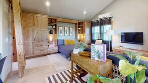 Uma sala de estar moderna com um sofá, uma mesa de centro e uma televisão. Há um armário de madeira e uma cama com cabeceira azul.