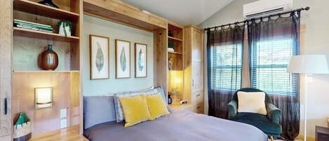 A bedroom with a large bed, wooden shelving, a desk, a chair, and a window with curtains.