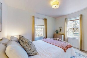 Ein Schlafzimmer mit einem Bett, zwei Vorhängen an den Fenstern, einem Nachttisch mit Pflanze und einer Wandleuchte.