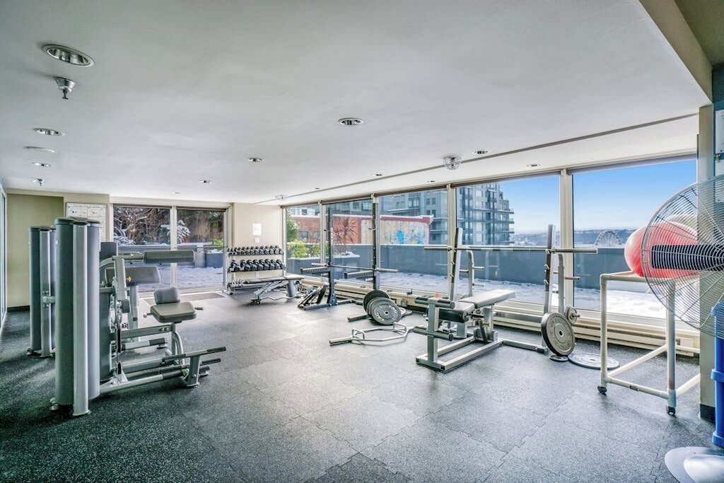 Un gimnasio con diferentes máquinas de ejercicio, amplias ventanas y vistas a la ciudad.