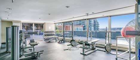 Un gimnasio con diferentes máquinas de ejercicio, amplias ventanas y vistas a la ciudad.