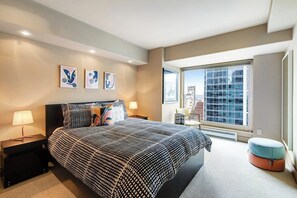 Une chambre moderne avec un grand lit, deux tables de chevet, une fenêtre donnant sur la ville et des œuvres d’art accrochées au mur.