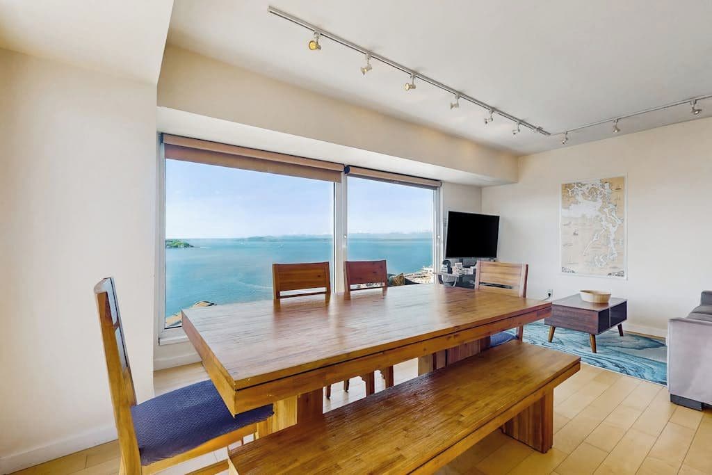 Un'area pranzo moderna con tavolo e sedie in legno, una TV a schermo piatto e un'ampia finestra vista mare.