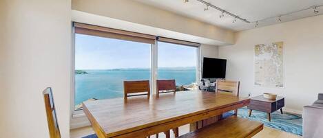 Un'area pranzo moderna con tavolo e sedie in legno, una TV a schermo piatto e un'ampia finestra vista mare.