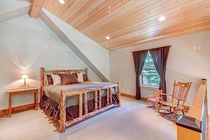 Ein Schlafzimmer mit schrägem Dach, einem hölzernen Bett, einem Nachttisch, einem Stuhl und einem Fenster mit Vorhängen.
