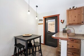 Eine kleine Küche mit schwarzer Arbeitsplatte, zwei Barhocker, einem Holzkorpus und einer Tür mit Blick auf ein Strohfeld.