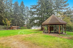 Ein Pavillon mit Dach und Bänken, umgeben von einer Rasenfläche und Bäumen.