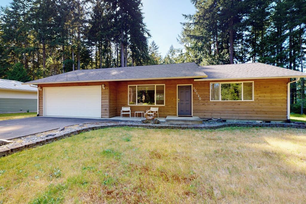 A single-story wooden house with a garage, a front porch with a table and chairs, and a small garden area.