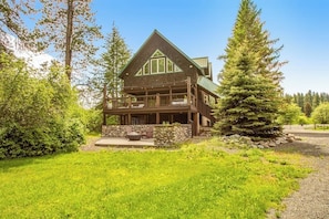A two-story lodge with a stone foundation, a covered porch, and a large evergreen tree in the foreground. - Quiet 2BR riverfront cabin with wood stove (Cle Elum)