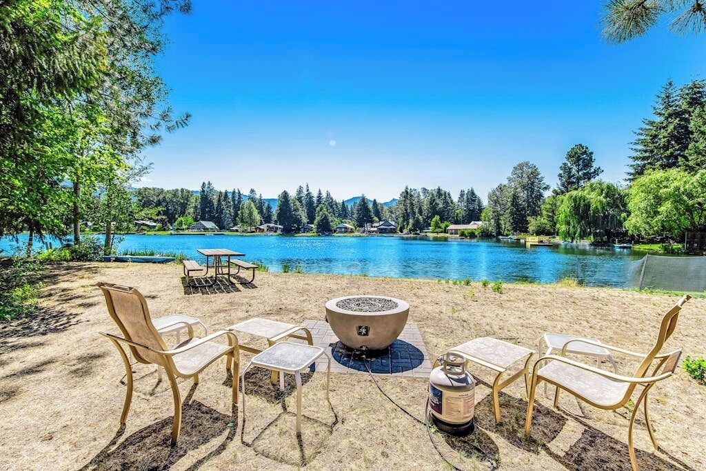 A lakeside outdoor seating area with a fire pit and chairs, surrounded by trees and a view of a lake.