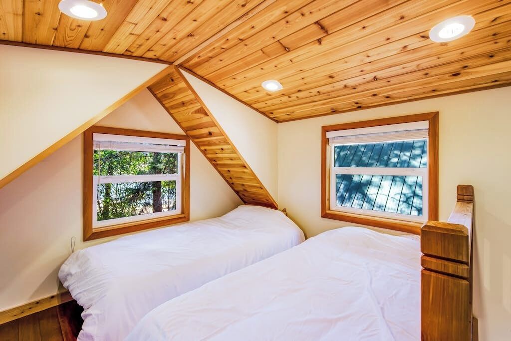 A bedroom with two beds, wooden ceiling beams, and windows with views of trees.