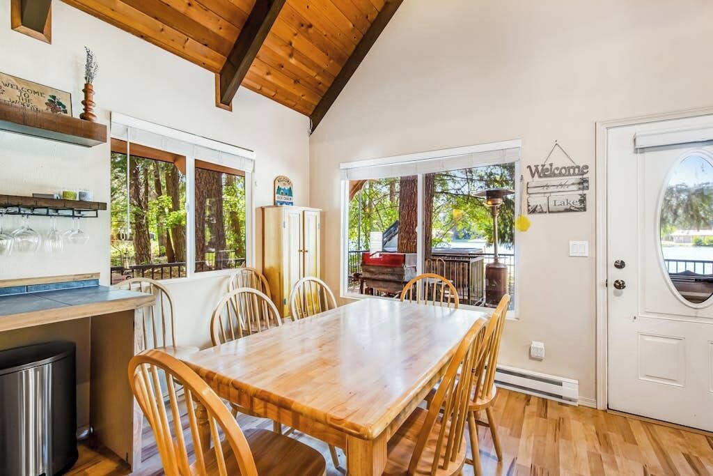 A dining area with a wooden table and chairs, a kitchen area with a refrigerator and a stove, and a view of the outdoors through large windows.