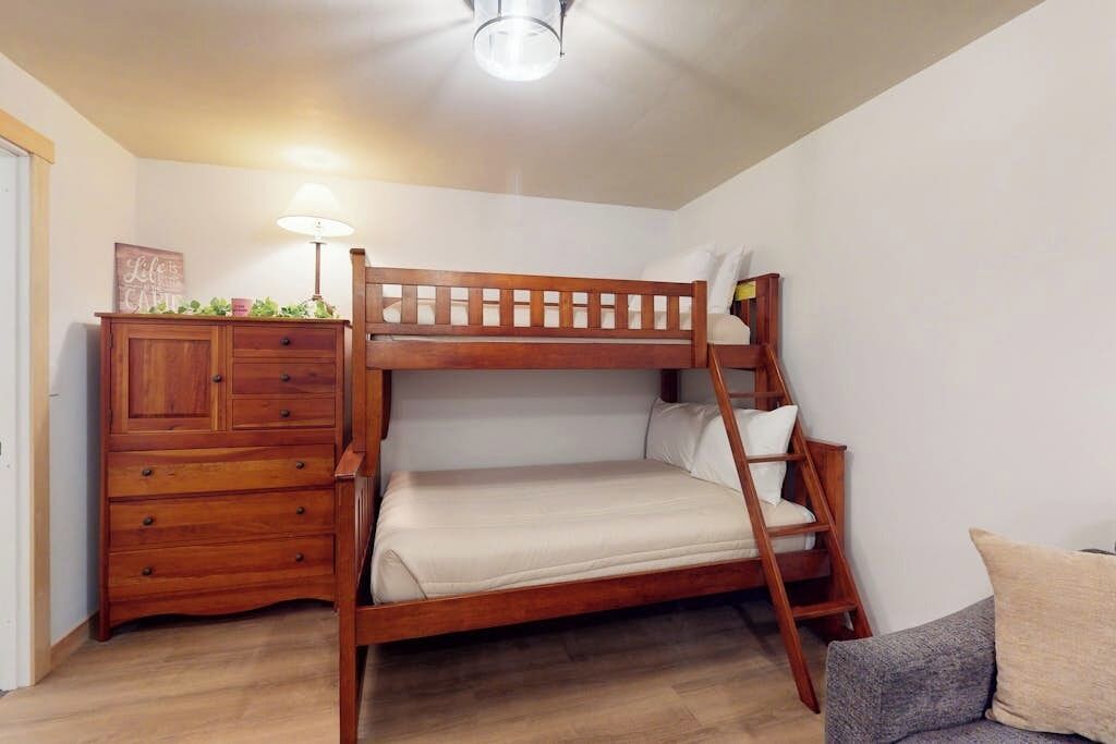 Una cama litera con estructura de madera, un aparador con cajones, una lámpara y un sofá gris en una habitación con suelo de madera y paredes blancas.