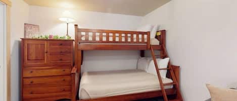Una cama litera con estructura de madera, un aparador con cajones, una lámpara y un sofá gris en una habitación con suelo de madera y paredes blancas.
