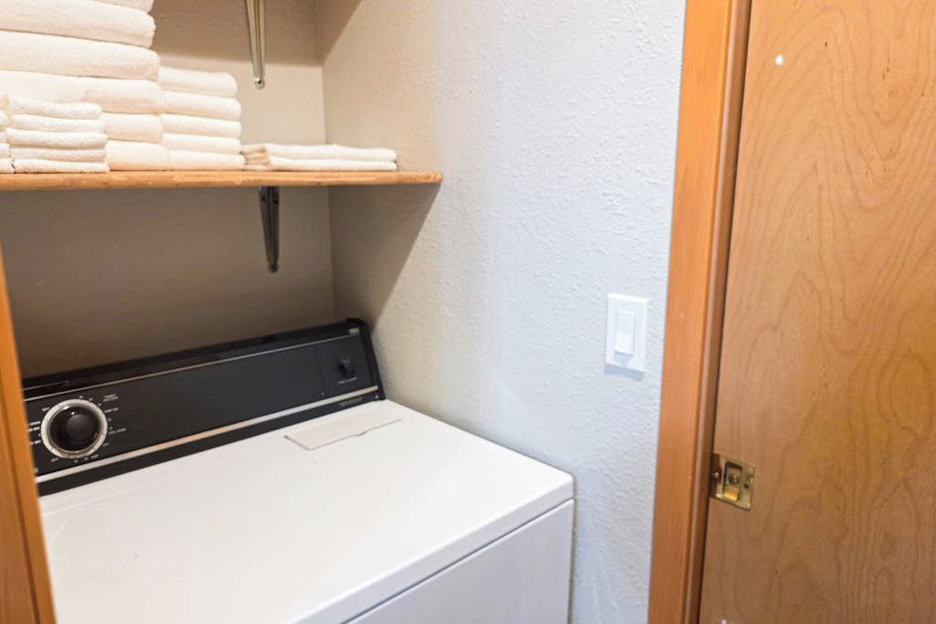 Un espace buanderie avec un lave-linge blanc, une étag ère où sont pliées des serviettes et une porte en bois.