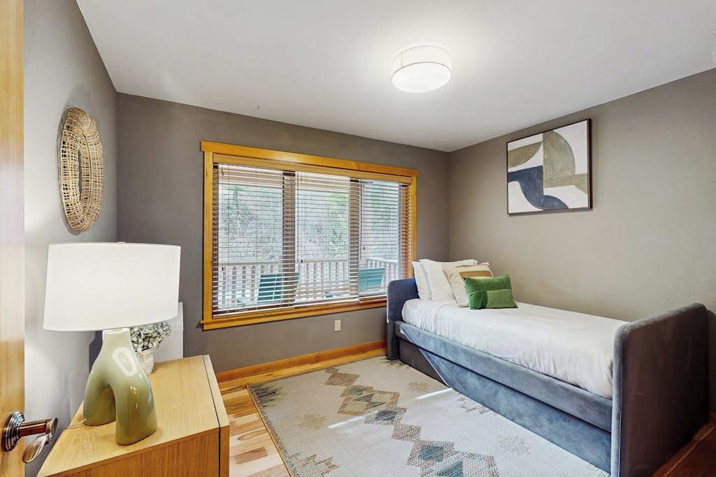 Une chambre à coucher avec un lit, une table de chevet avec une lampe, une fenêtre avec des stores et une œuvre d’art accrochée au mur.