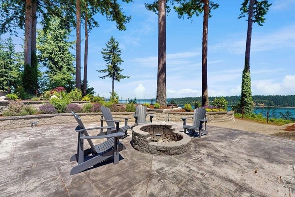 Een terras met Adirondack-stoelen en een vuurkorf van natuursteen, omringd door hoge bomen en uitzicht op een meer.