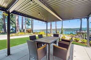 Um pátio externo com área de estar coberta, uma mesa e vista para um lago.