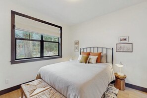Ein Schlafzimmer mit großem Fenster, einem Bett mit vielen Kissen, einem Nachttisch mit Lampe und einer kleinen Bank.