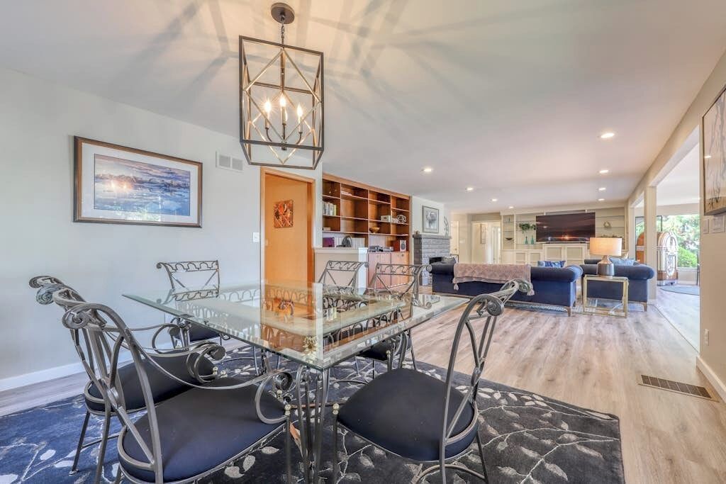A dining area with a glass table and metal chairs, a bookshelf, and a sofa in the background.