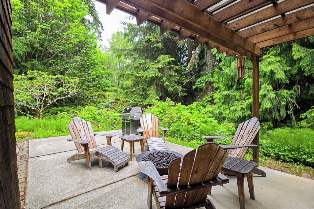 A covered patio with Adirondack chairs, a fire pit, and a grill, surrounded by lush greenery.