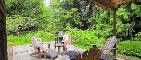A covered patio with Adirondack chairs, a fire pit, and a grill, surrounded by lush greenery.