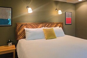 Ein ordentlich bezogenes Bett mit einem Holzkopfteil, zwei Wandleuchten und zwei gerahmten Kunstwerken an den Wänden.