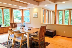 Ein Essbereich mit Holztisch und -stühlen, großen Fenstern mit Blick auf Grünflächen und einem gerahmten Bild an der Wand.