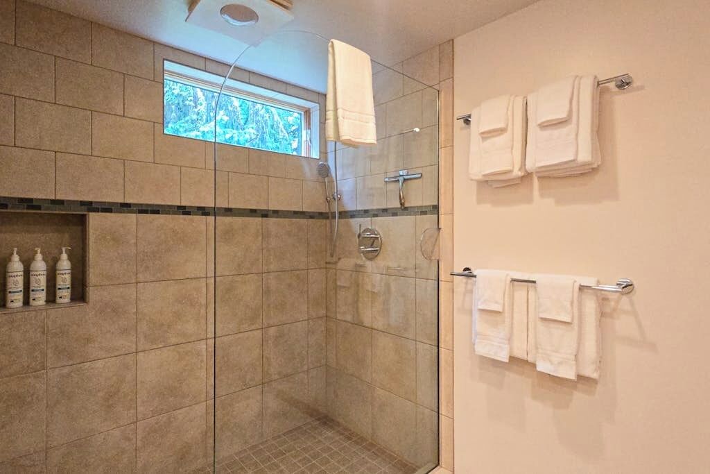 Casa de banho moderna com cabine de duche em vidro, azulejos bege, uma pequena janela e toalhas bem organizadas.