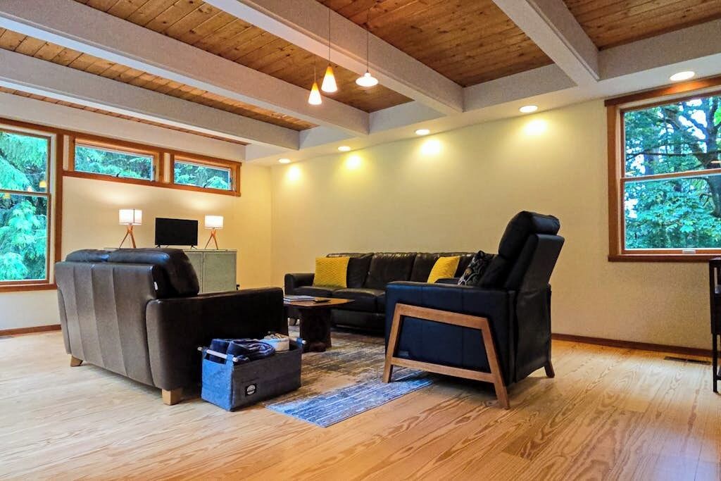 A living room with a sofa, armchair, and coffee table, wooden beams on the ceiling, and large windows with a view of trees.