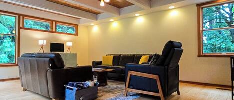Una sala de estar con sofá, sillón y mesa de centro, vigas de madera en el techo y grandes ventanales con vistas a árboles.