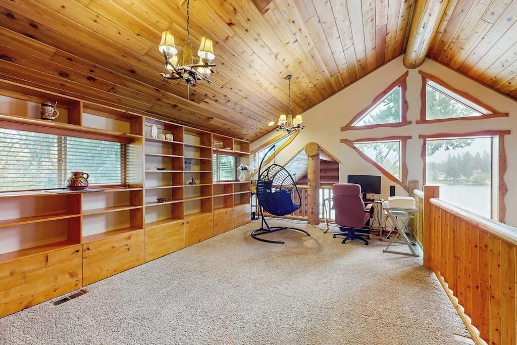 A wooden interior with a sloped ceiling, built-in shelves, a hanging chair, a desk with a computer, and large windows.