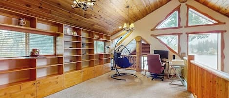 A wooden interior with a sloped ceiling, built-in shelves, a hanging chair, a desk with a computer, and large windows.