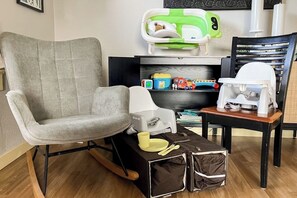 Ein Zimmer mit grauem Sitzmöbel, einem kleinen Tisch mit Babysitz, einer Wickelstation und einem Regal mit Spielzeug.