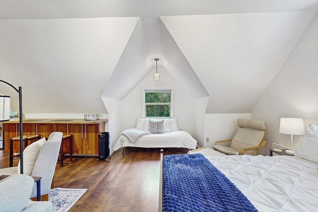 A bedroom with a loft-style ceiling, a bed with a blue blanket, a chair, a desk, and a window.