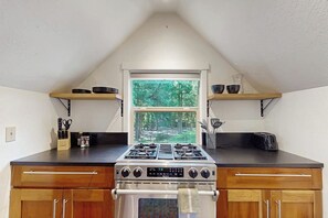 A kitchen with a gas stove, oven, and a window with a view of trees. - 0BR Studio on lush grounds with nearby horses (Bainbridge Island)