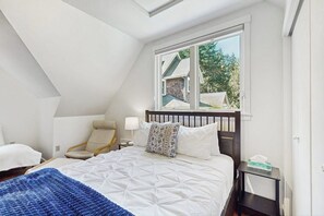 A bedroom with a bed, a chair, a nightstand, and a window with a view of a house. - 0BR Studio on lush grounds with nearby horses (Bainbridge Island)