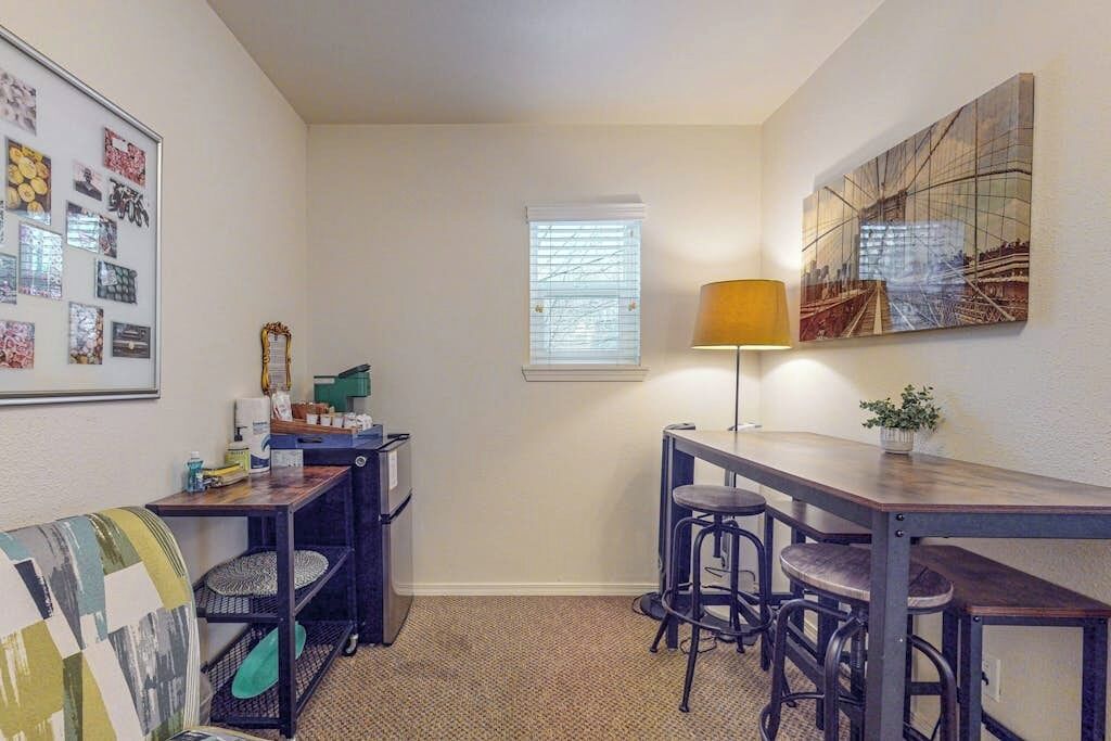 A compact room with a desk, two bar stools, a small table, a lamp, a window with blinds, and a wall-mounted shelf with various items.