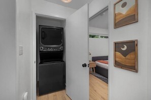 A compact kitchen with a microwave and refrigerator, a bedroom with a bed and nightstand, and two framed artworks on the wall. - 3BR waterfront home with patio & wildlife watching (Lilliwaup)