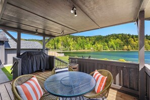 A covered outdoor deck with a glass table and wicker chairs, overlooking a lake and forest. - 3BR waterfront home with patio & wildlife watching (Lilliwaup)