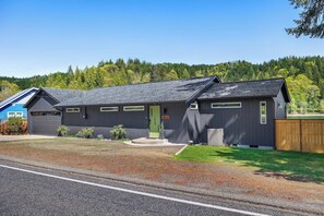 A two-story building with a green door, a covered outdoor area, and a view of a forested hill in the background. - 3BR waterfront home with patio & wildlife watching (Lilliwaup)