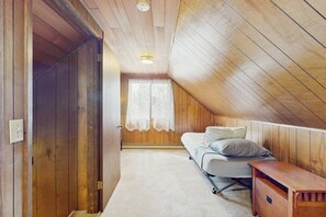 Ein Zimmer mit schrägen Wänden, einem Bett mit Kissen, einer Holzbank und einer Tür zu einem weiteren Raum.