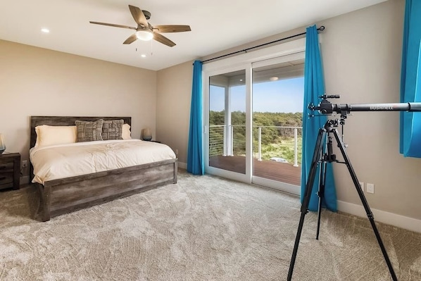 A bedroom with a bed, a ceiling fan, a balcony with blue curtains, and a telescope on a tripod.